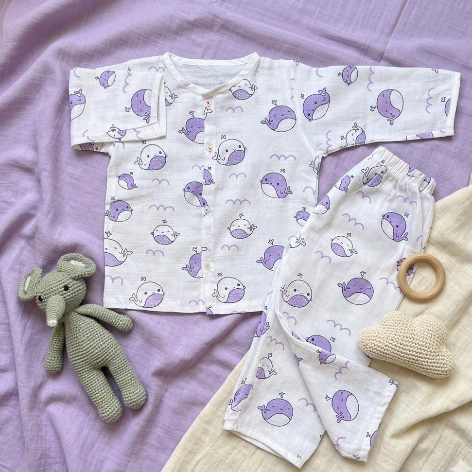 Baby outfit with purple whale pattern on a purple blanket, accompanied by a green toy elephant and wooden teething ring.