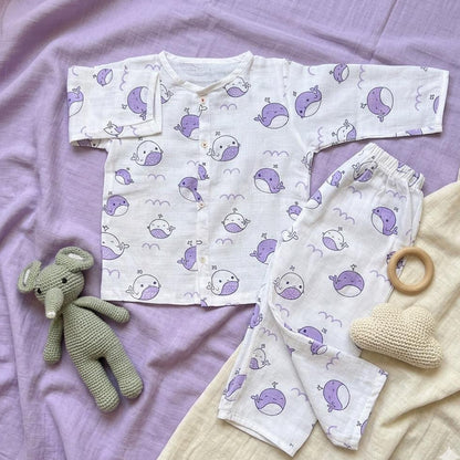 Baby outfit with purple whale pattern on a purple blanket, accompanied by a green toy elephant and wooden teething ring.